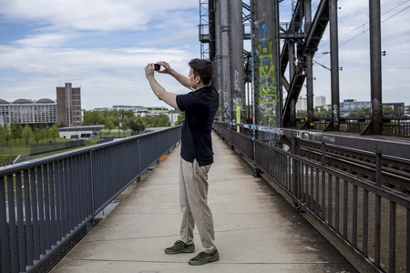 A man taking a photograph of a city with a smartphone while standing on a bridge walkway with rail...