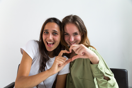 Two beautiful young women looking at the camera and making a heart shape hand gesture. Medium shot.の写真素材