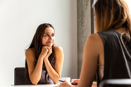 Female colleagues talking in the office seated across work desk. Medium shot.の写真素材
