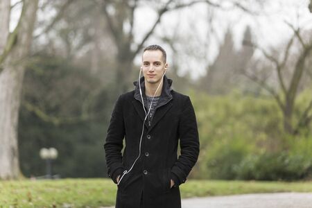 Young Caucasian Man Listening to Earphones and Wearing Black Coat Outdoorsの写真素材