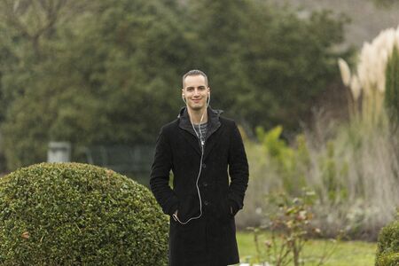 Young Man Wearing Coat and Listening to Earphones Posing in Gardenの写真素材