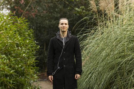 Young Man Posing in Garden Wearing Coat and Listening to Headphonesの写真素材