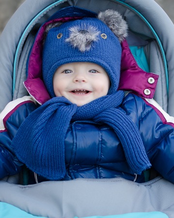 Happy baby boy sitting in a stroller for a walk in winter outdoor. Adorable smiling baby boy in blue winter clothes. Portrait of a cute baby with huge blue eyes in buggy outdoors on walking.の写真素材