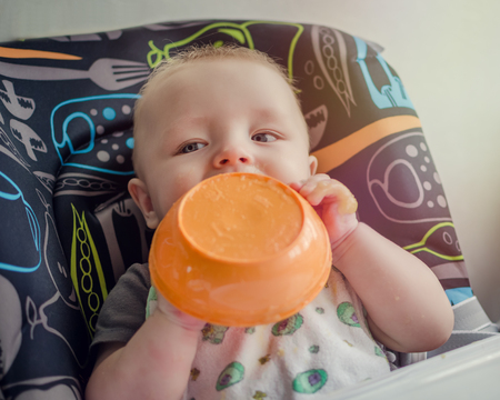 Baby eating in high chair from the plate, holding it in the hands. Baby boy feeding himself with messy face. Child having breakfast, making mess. Baby at meal time. Baby's first solid food. Baby food.の写真素材