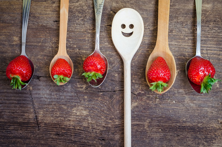 Strawberries on vintage spoons and wooden spoon with smiley face over old rustic wooden table top. Bright positive background with smileys. Summer concept for summer season. Top view.の写真素材