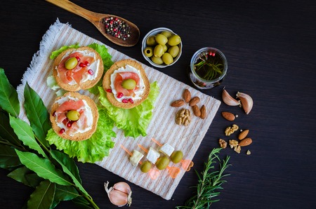 Delicious bruschetta with prosciutto, olives and pomegranate on dark background with olive oil with spices, rosemary, Bay leaf, garlic, nuts and wooden spoon with pepper. Italian appetizer. Top view.の写真素材