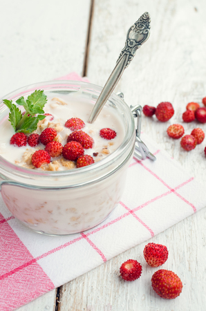 Fresh milk yoghurt with wild strawberries and granola served in a glass jar with mint leaves on white wooden table with striped red napkin. Healthy summer breakfast concept. Selective focus.の写真素材