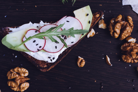 Sandwiches with avocado, radish, arugula, cheese and nuts on a rye bread with sunflower seeds for healthy breakfast on dark wooden background. Vegetarian food. Top view.の写真素材