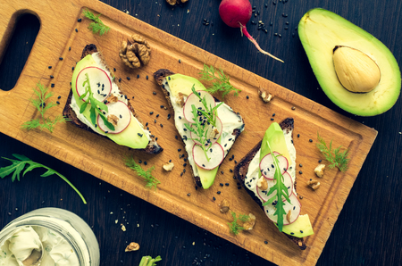Sandwiches with avocado, radish, arugula, cheese and nuts on a rye bread with sunflower seeds on wooden board on dark background. Healthy breakfast. Vegetarian food. Top view.の写真素材