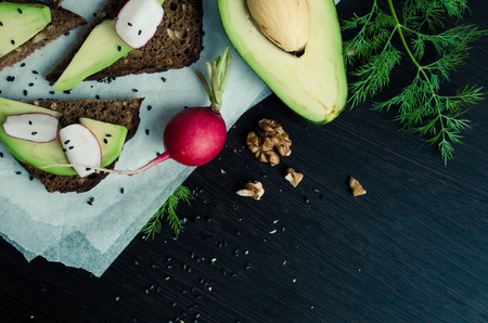 Sandwiches with avocado, radish, arugula, cheese and nuts on a rye bread with sunflower seeds for healthy breakfast on dark wooden background. Vegetarian food. Top view. Copy space.の写真素材
