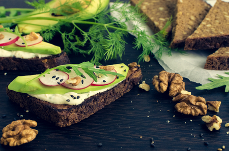 Sandwiches with avocado, radish, arugula, cheese, black sesame and nuts on a rye bread with sunflower seeds for healthy breakfast on dark wooden background. Vegetarian food. Selective focus.の写真素材
