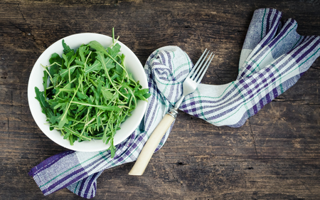Fresh arugula in a plate with a fork on rustic wooden background with place for text. Healthy organic diet vegan food concept. Green meal. Top view. Copy space.の写真素材