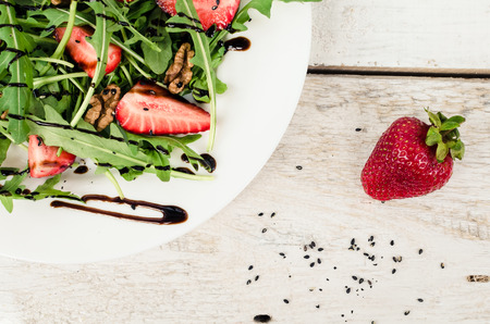 Fresh salad with arugula, strawberries, nuts, black sesame seeds and balsamic glasse sauce served on white plate on rustic wooden table. Healthy organic diet food concept. Top view.の写真素材