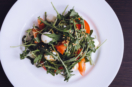 Fresh summer salad with tomatoes, fresh arugula leaves, mini mozzarella cheese and sesame seeds on white plate over dark wooden baclground. Healthy diet food concept. Top view.の写真素材