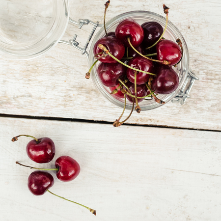 Red ripe juicy cherry in glass jar on white rustic wooden background. Sweet summer berries. Freshly harvested merry. Directly above. Top view. Copy space. Square.の写真素材