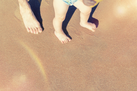 Toddler baby doing his first steps on the beach. Bare feet father and son staying on the sand near the bank in sunny day. Summertime holidays concept. Top view. Copy space.の写真素材