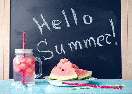 Hello summer written on the blackboard with drink in glass mason jar with slices of watermelon and ice on blue background. Fresh fruit flavored infused detox water. Start Summer Concept.の写真素材