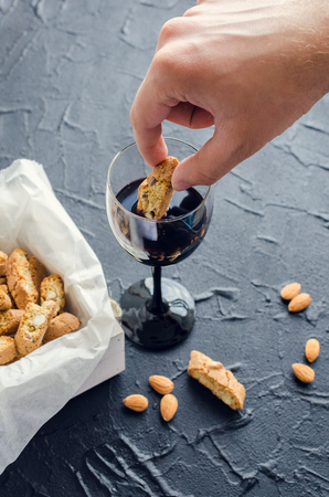 Male hand dipping traditional italian almond cookies cantuccini into red wine on black stone background. Homemade biscotti on dark concrete table.の写真素材