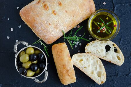 Sliced freshly baked loaf of traditional italian bread ciabatta on black stone table with rosemary, salt, olive oil and olives. Italian food concept. Top view.の写真素材