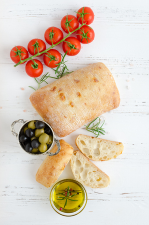 Sliced freshly baked loaf of traditional italian bread ciabatta on white rustic wooden table with rosemary, olive oil, tomatoes and olives. Italian food concept. Top view.の写真素材