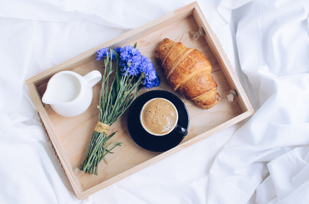 Romantic summer breakfast in bed, tray with fresh croissant, cup of coffee espresso with milk and bouquet of blue cornflowers. Good morning concept. Enjoy slow life. Top view.の写真素材