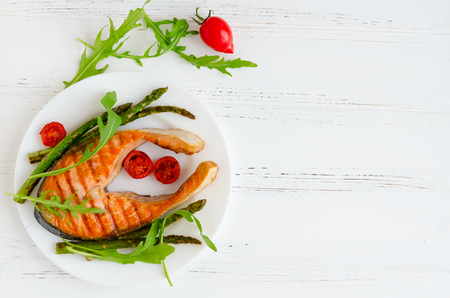 Grilled salmon steak with asparagus and tomatoes cherry on a plate with arugula on white wooden background with place for text. Healthy lunch concept. Top view. Copy space.の写真素材