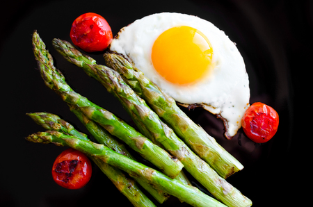 Fried egg and roasted fresh asparagus with tomatoes cherry in black plate. Healthy lunch concept. Delicious, nutritious eating. Top view.の写真素材
