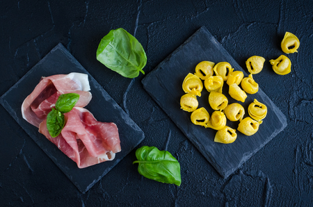 Freshly prepared raw homemade meat prosciutto filled tortellini ready to cook on dark stone plate with basil leaves on black background. Traditional Italian cuisine concept. Top view.の写真素材