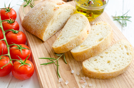 Sliced freshly baked ciabatta bread on wooden cutting board on white rustic table with rosemary, salt, tomatoes and olive oil. Italian food concept.の写真素材