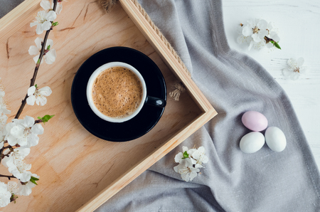 Cup of coffee espresso, colorful chocolate eggs and cherry blossom on wooden tray on white shabby chic table with gray tablecloth. Easter treats concept. Top view.の写真素材