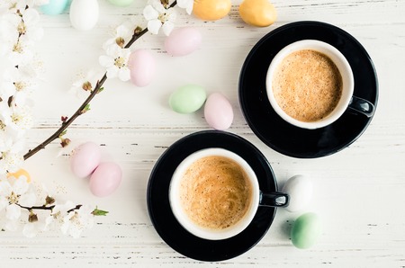 Two cups of coffee espresso, colorful chocolate eggs and cherry blossom on white shabby chic background. Easter treats concept. Top view.の写真素材