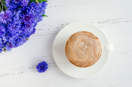 A cup of morning drink cappuccino with whipped cream and bouquet of blue cornflowers on white wooden table. Top view.の写真素材