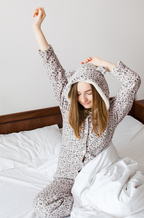 Happy young woman enjoying sunny morning. Caucasian girl in cute warm pajamas stretching in the bed and smiling after waking up. Good start of the day concept.の写真素材