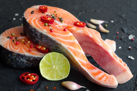 Two fresh raw salmon steaks with pepper corns, chili, salt, lemon and garlic on blackの写真素材