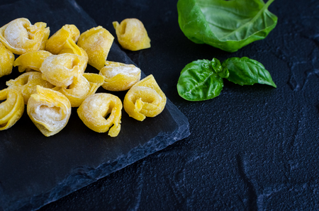 Freshly prepared homemade raw flouring tortellini ready to cook on dark stone plate with basil leaves on black background. Traditional Italian cuisine concept. Copy space.の写真素材