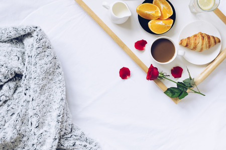 Romantic Valentines Day breakfast in bed, tray with fresh croissant, cup of coffee espresso with milk, fresh oranges and red rose.の写真素材