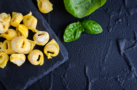 Freshly prepared homemade raw flouring tortellini ready to cook on dark stone plate with basil leaves on black background. Traditional Italian cuisine concept. Top view. Copy space.の写真素材
