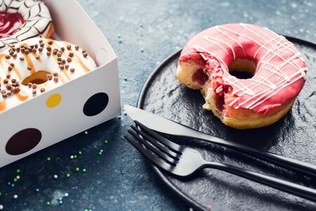 Pink frosted donut with bite missing on a plate with assortment donuts box on dark background. Top...