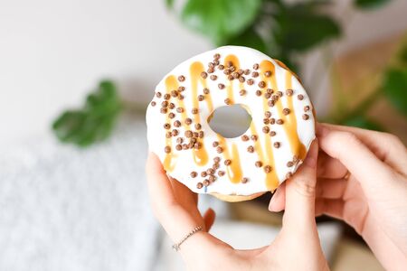 Female hand holding delicious donut with white glaze, caramel and chocolate sprinkles. Party concept. No diet. Candy bar. Proper nutrition or sweets, dessert fast food, dieting morning concept.の写真素材