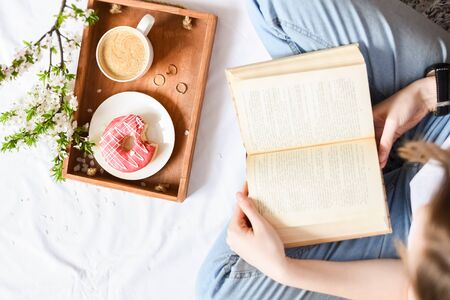 Spring breakfast in bed. Tray with strawberry pink donut with coffee and spring blossom. Girl reading a book. Top view. Copy space.の写真素材