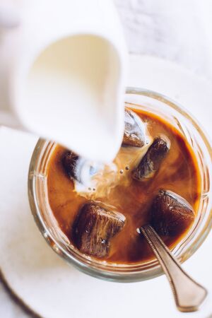 Pouring milk from a jug to a glass of espresso ice cubes on white  with copy spaceの写真素材
