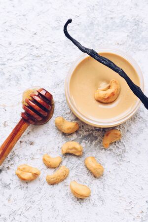 Cashew nut butter in glass jar. Homemade raw organic cashew nuts paste on grey background with honey and vanilla. Healthy natural food concept. Top view.の写真素材
