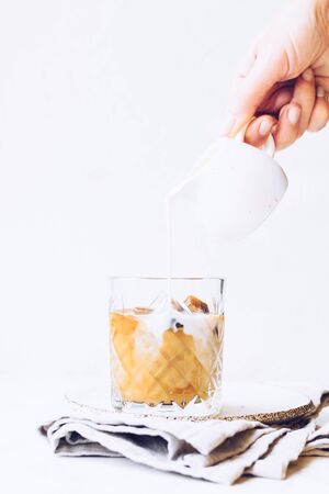 Pouring milk from a jug to a glass of espresso ice cubes on whiteの写真素材