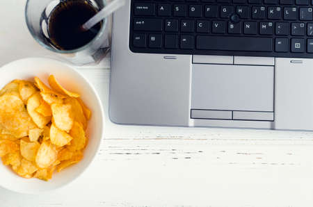 Working at home and snacks. Home office with unhealthy lifestyle. Leisure with laptop and fast food. Potato chips, cola and computer on the table. Top view.の写真素材