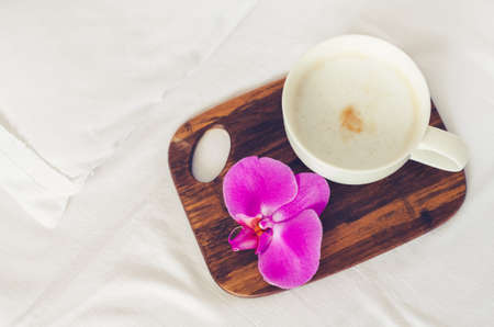 Cup of coffee cappuccino with flower on wooden board on a bed on cozy lazy sunday. White bedding sheet, blanket and pillows. Good morning concept. Enjoy slow life. Top view.の写真素材