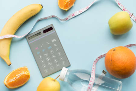 Calculator with numbers 1200, bottle of water, measuring tape and fruits on blue background. Healthy eating concept - calculate daily nutrition intake. Top view. Copy space.の写真素材