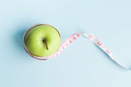 Apple with measuring tape on blue background. Weight loss, counting calories and healthy eating concept - calculate daily nutrition intake. Top view. Copy space.の写真素材