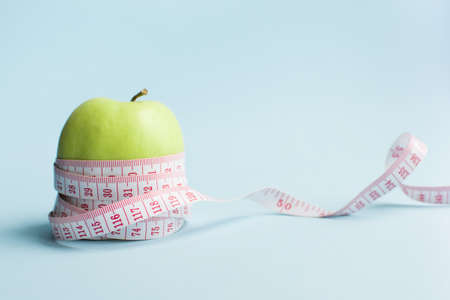 Apple with measuring tape on blue background. Weight loss, counting calories and healthy eating concept - calculate daily nutrition intake. Copy space.の写真素材