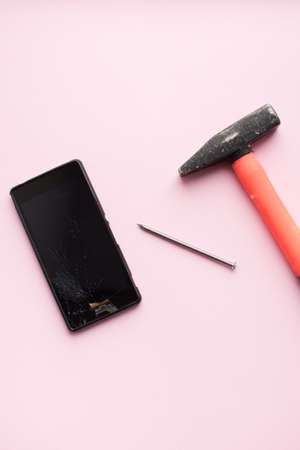 Hammer, nail and mobile phone with broken display on pink background. Crashed smartphone concept....