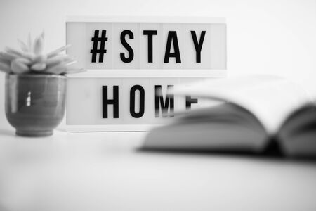 Sign Stay Home, home plant and open book in soft focus on white background. Black and white. Concept...